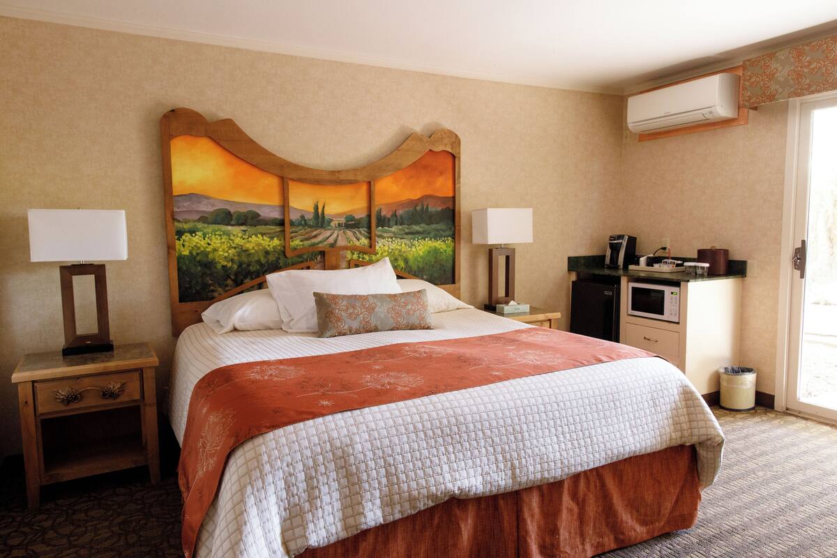 A hotel room with a large bed, two bedside tables with lamps, a desk with a microwave, and an air conditioning unit.