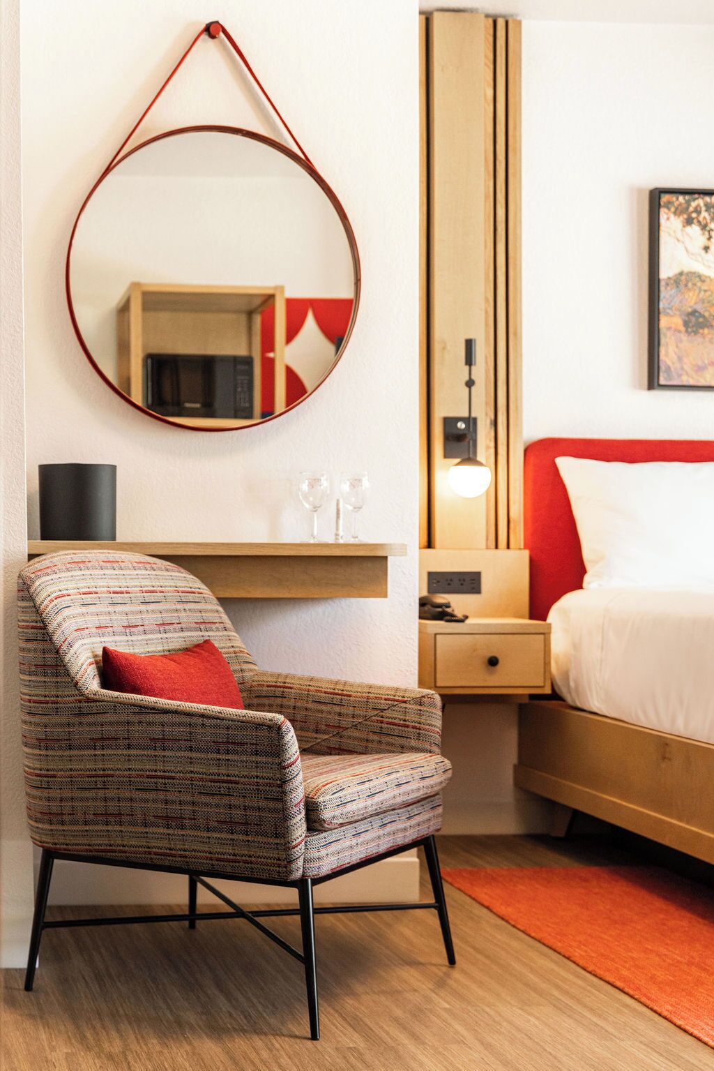 A hotel room with a round mirror, a red and white bed, a wooden nightstand, a small table with glasses, and a patterned armchair.