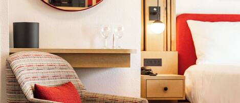 A hotel room with a round mirror, a red and white bed, a wooden nightstand, a small table with glasses, and a patterned armchair.