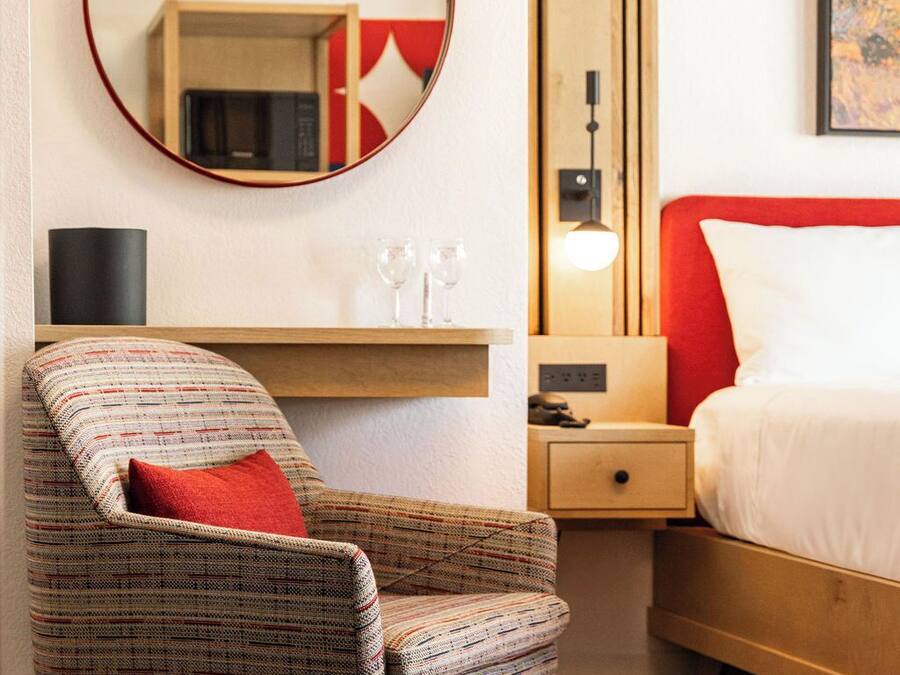 Une chambre d’hôtel avec un miroir rond, un lit rouge et blanc, une table de chevet en bois, une petite table avec des verres et un fauteuil à motifs.