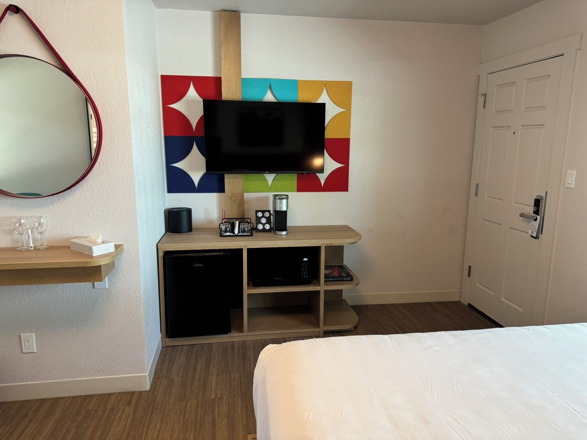 A hotel room with a bed, a TV mounted on a colorful wall, a small desk with a coffee maker, and a minibar.