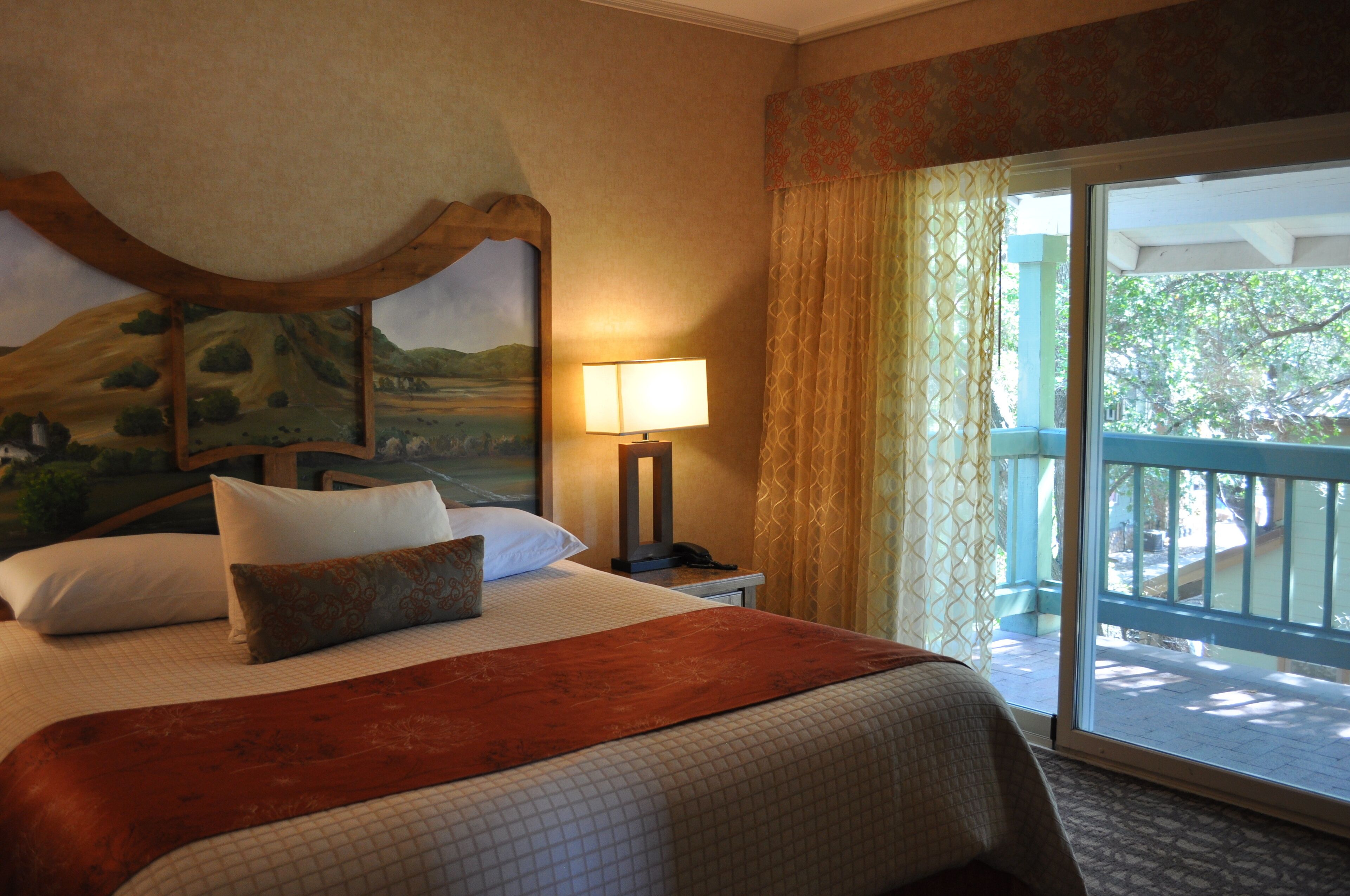 Quarto de hotel com uma cama, mesa de cabeceira, luminária, quadro e vista para o exterior por uma porta deslizante.