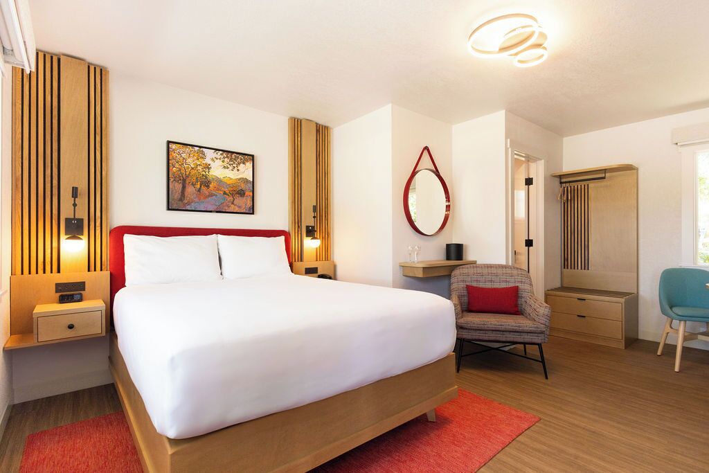 A modern hotel room with a large bed, a bedside table with a lamp, a chair, a mirror, and a painting on the wall.