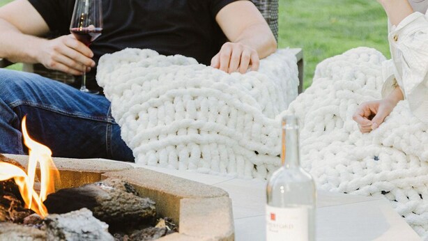 A couple sitting on wicker chairs with a fire pit in front, holding wine glasses.