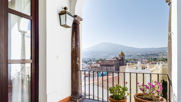 Uma varanda com azulejos de terracota, um vaso de plantas e vista para uma cidade com uma igreja e montanhas ao longe.