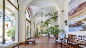 Um hall amplo com tetos altos, portas em arco e chão de azulejos. Há vários bancos e plantas em vasos.