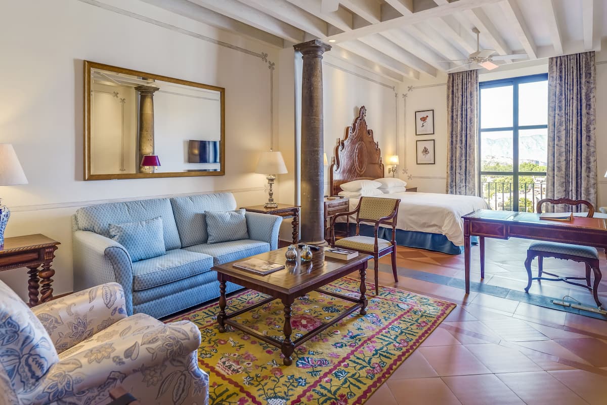 Habitación de hotel con una cama grande, zona de estar con sofá y sillones, una mesa de centro de madera, y vistas a montañas a través de la ventana.