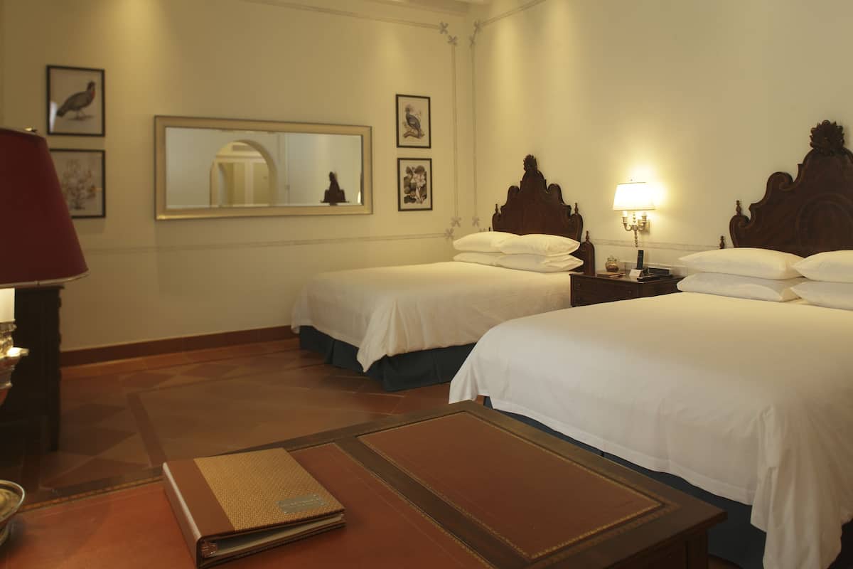 Habitación de hotel con dos camas, una mesita de noche con lámpara, un espejo y cuadros en la pared.
