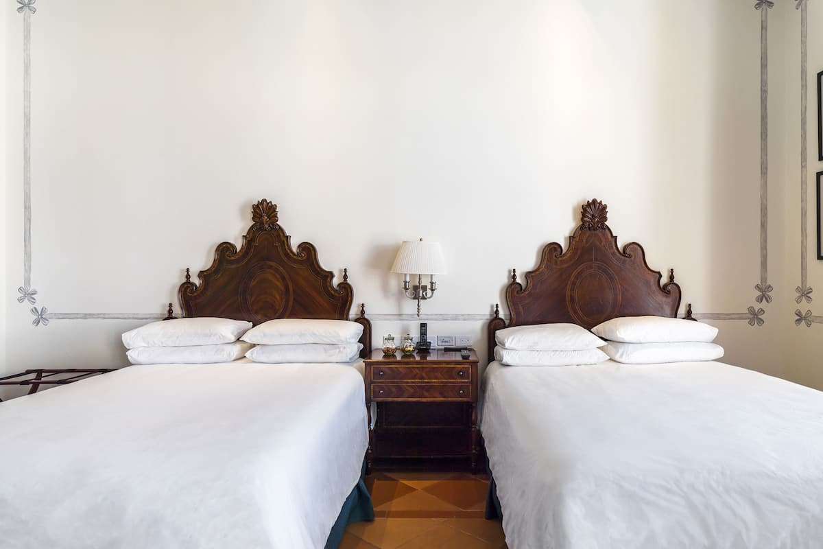 Habitación de hotel con dos camas, una mesita de noche, una lámpara y una pared con motivos florales decorativos.