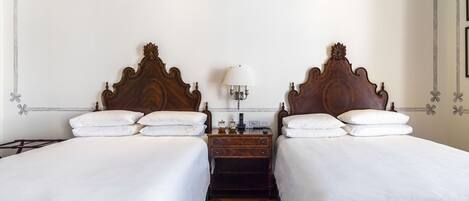 Quarto de hotel com duas camas, uma mesa de cabeceira, uma luminária e uma parede com padrões florais decorativos.