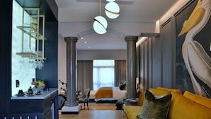 Une chambre d’hôtel avec un lit, un canapé jaune, un coin bar avec des verres et un grand tableau représentant un oiseau.