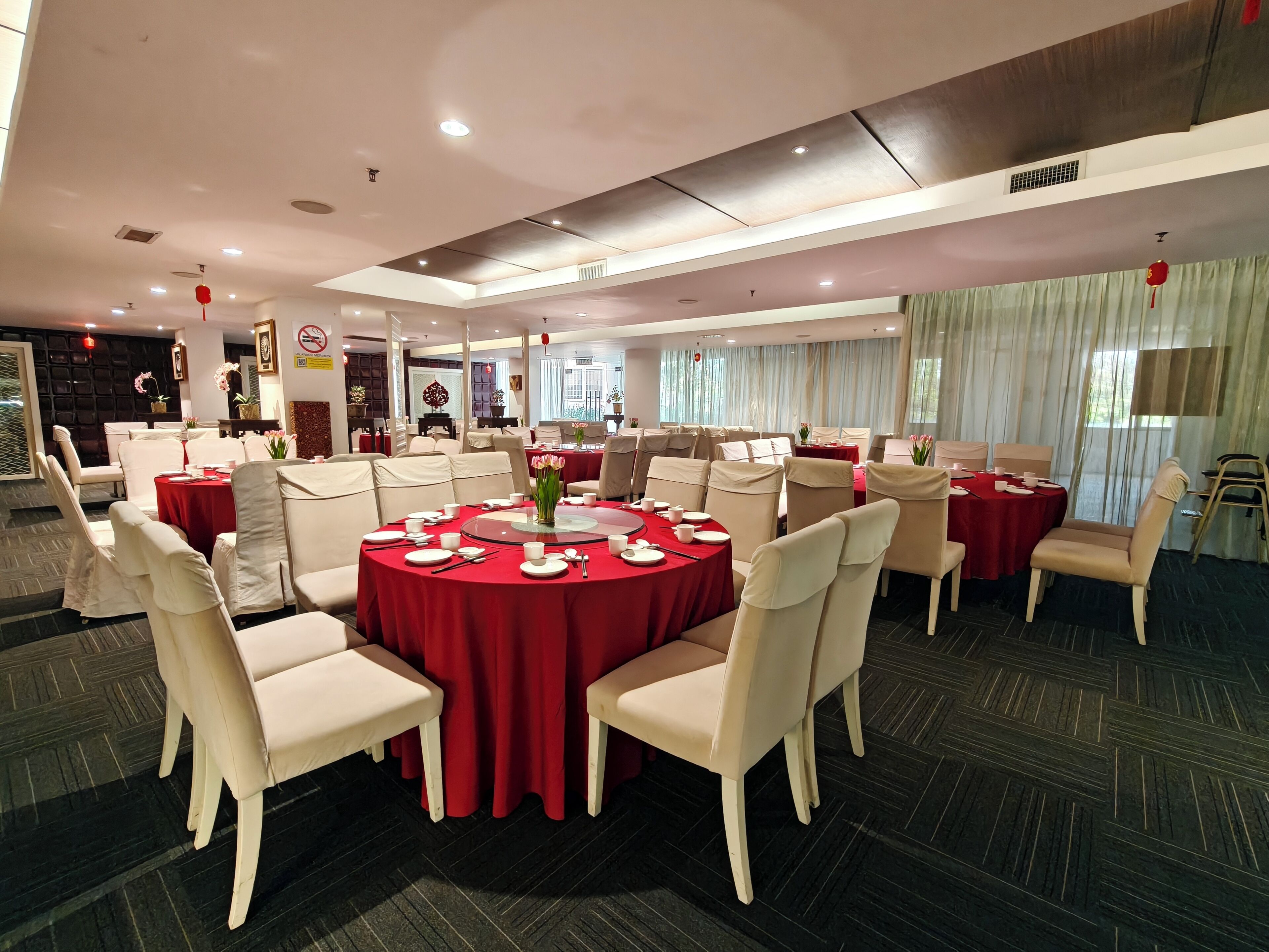 Un amplio salón de banquetes con varias mesas redondas, cada una con sillas blancas y manteles rojos.