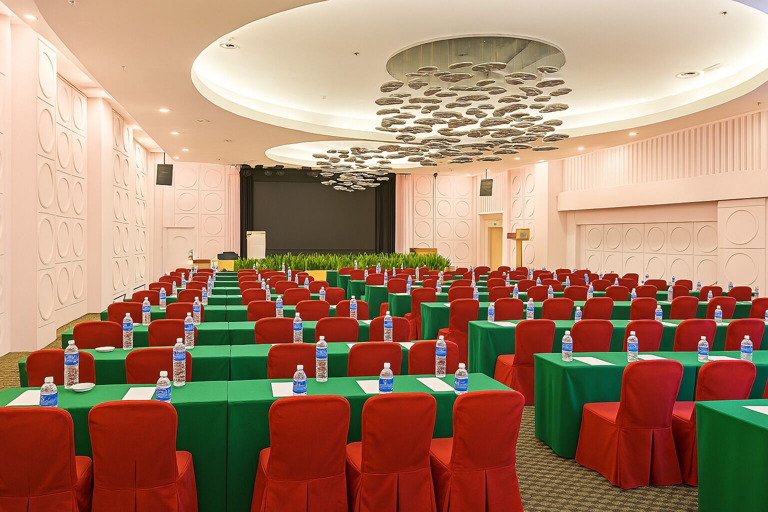 Ein Konferenzraum mit Tischen, Stühlen in Reihen, einer Bühne und einer großen Chandelier.