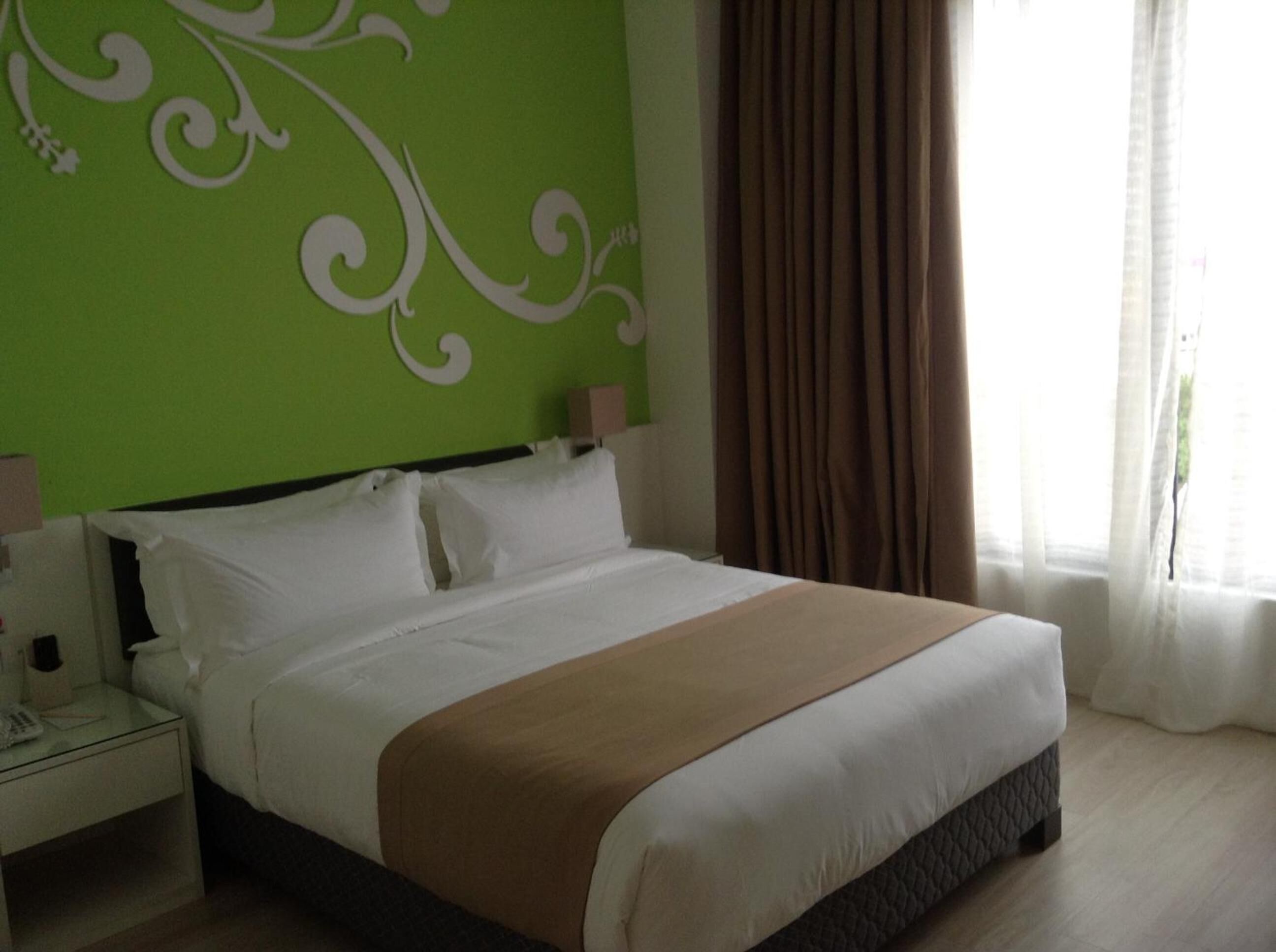 A neatly made bed with white linens and a brown throw, a bedside table with a lamp, and a green wall with decorative swirls.