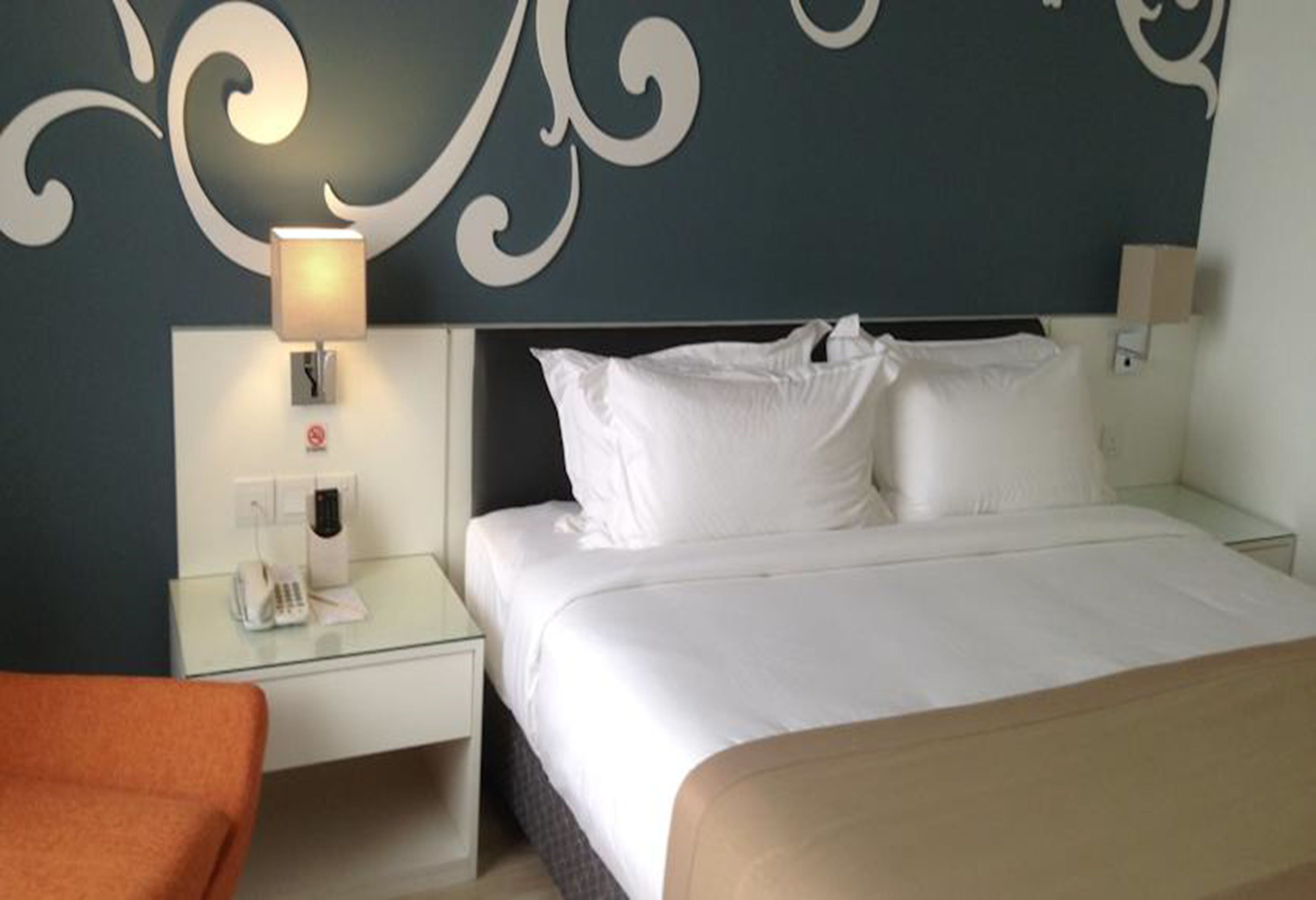A hotel room with a bed, a nightstand with a lamp, a phone, and a wall with a decorative design.