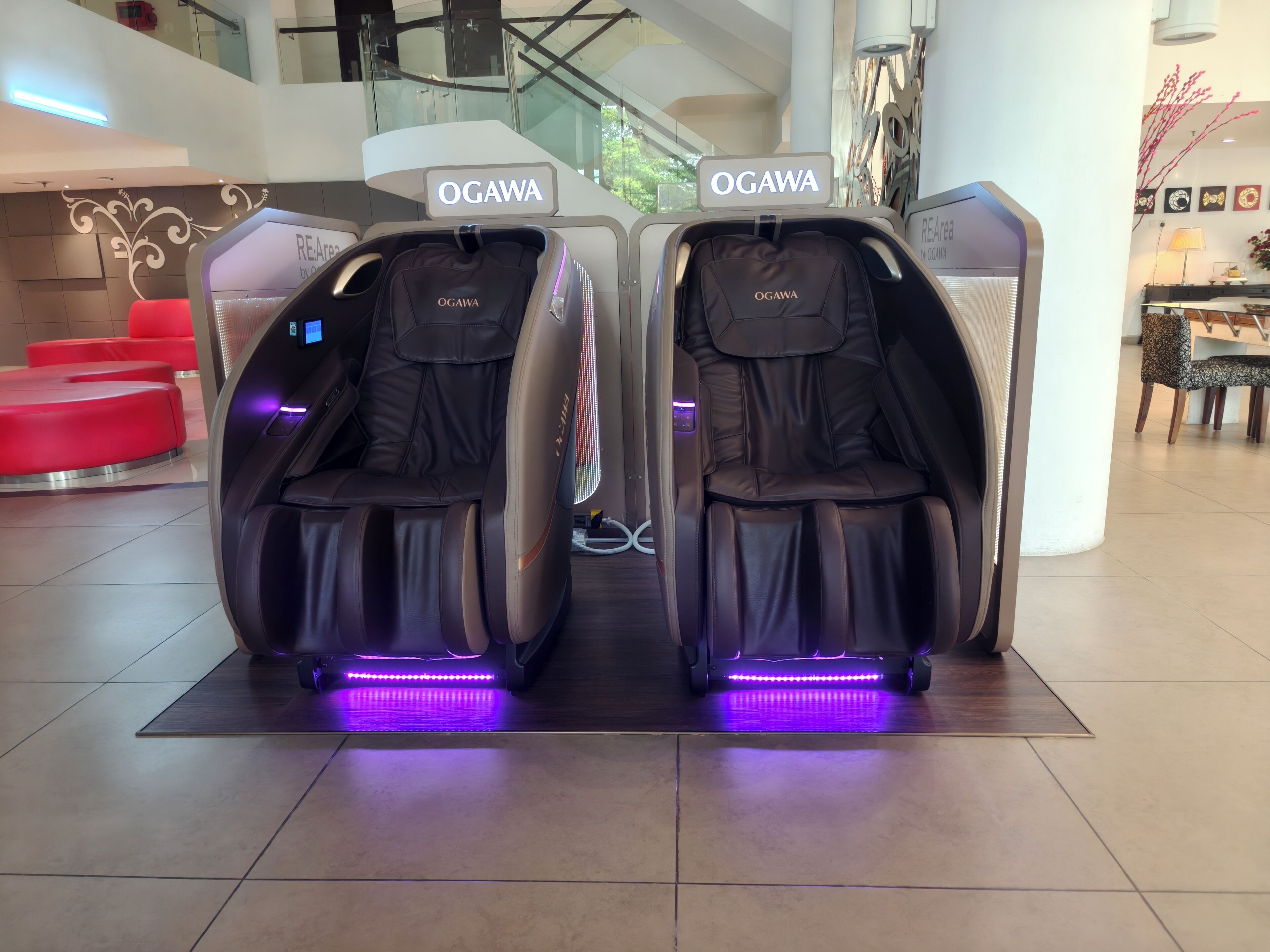 Two massage chairs labeled 'OGAWA' in a modern lobby with a glass staircase and red seating in the background.