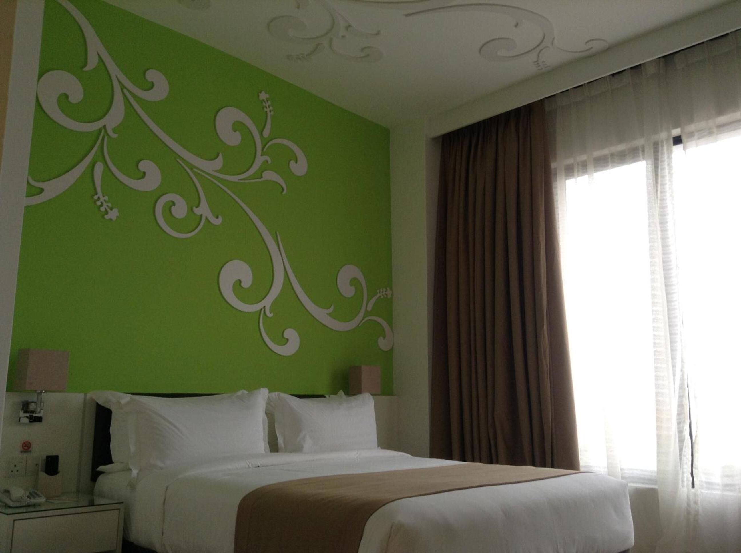 A hotel room with a green accent wall, a bed with white linens, a nightstand with a phone, and a window with curtains.