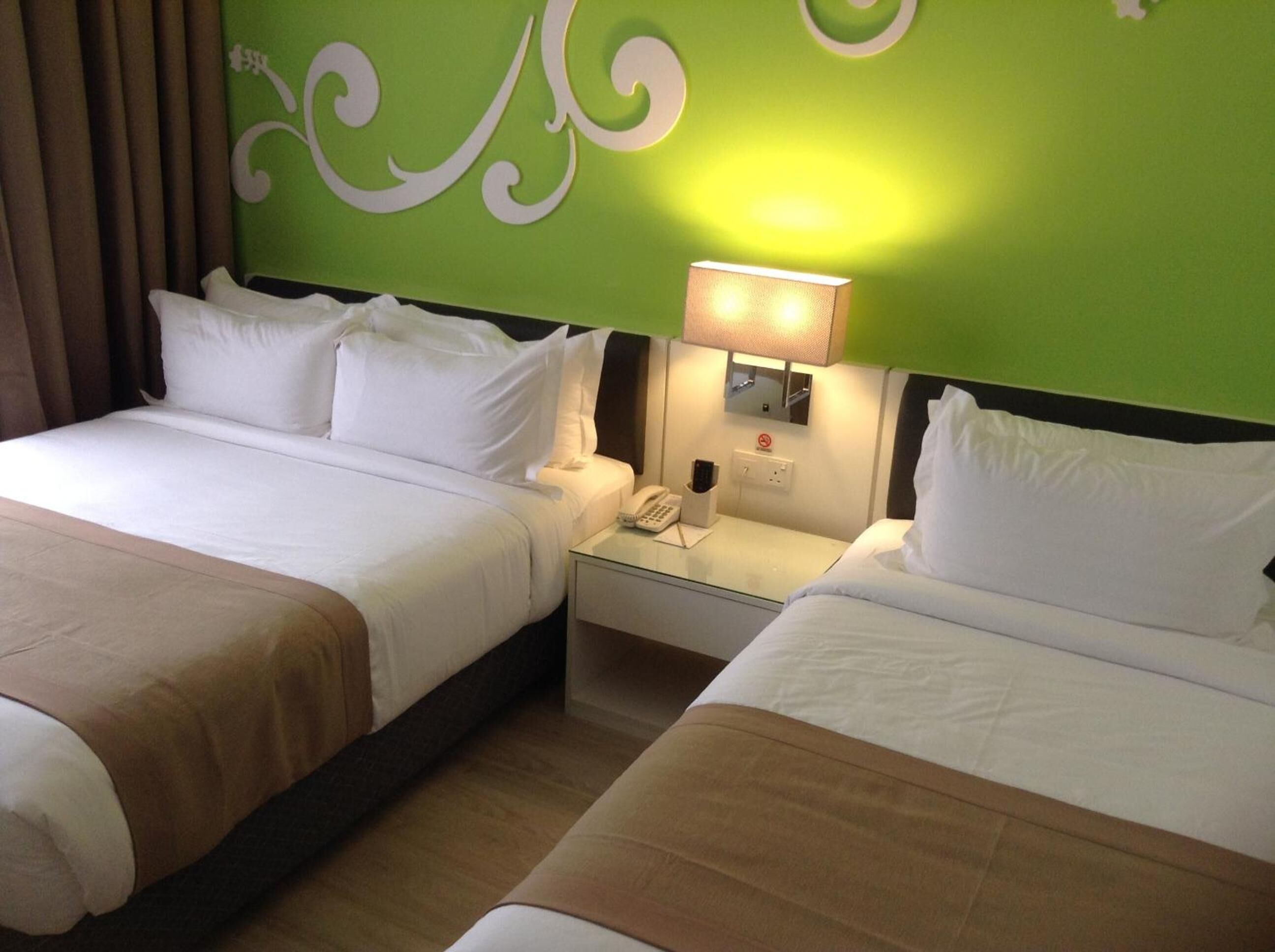 A hotel room with two beds, each with white linens and brown headboards. There is a nightstand with a lamp and a phone between the beds. The wall behind the beds is green with white decorative swirls.