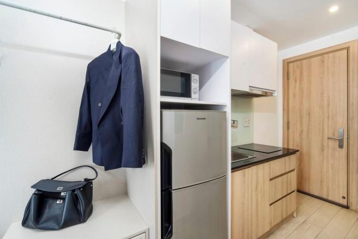 A compact kitchen with a refrigerator, microwave, and wooden cabinets. A coat is hanging on a rack, and a black bag is on the counter.
