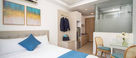 A hotel room with a bed, a desk with two chairs, a mirror, a kitchenette, and a shower area.