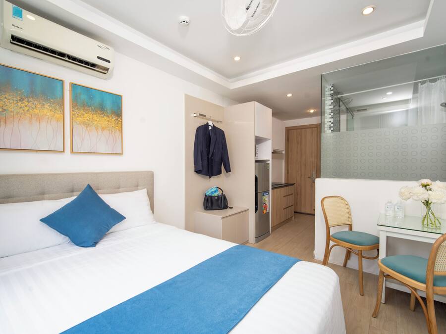 Quarto de hotel com cama, escrivaninha com duas cadeiras, espelho, cozinha compacta e área de chuveiro.