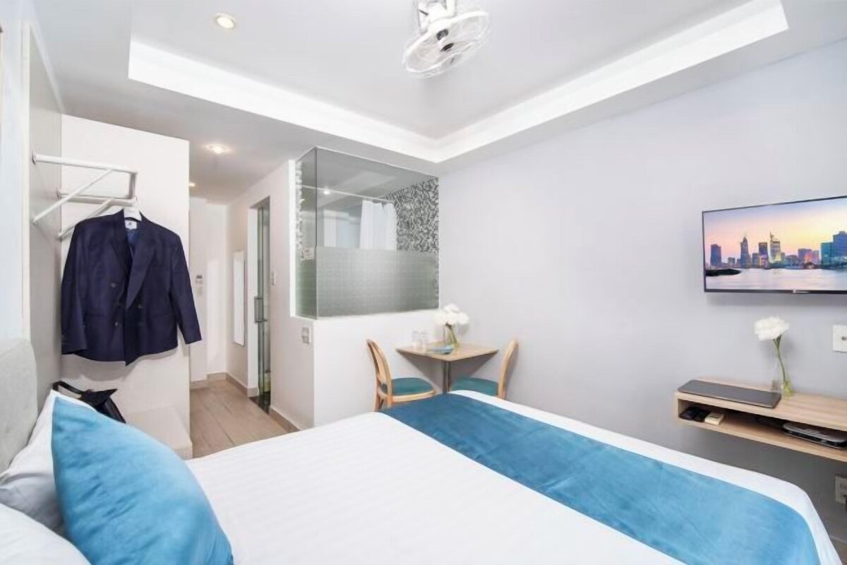 A modern hotel room with a bed, a desk, a chair, a TV, and a cityscape view.