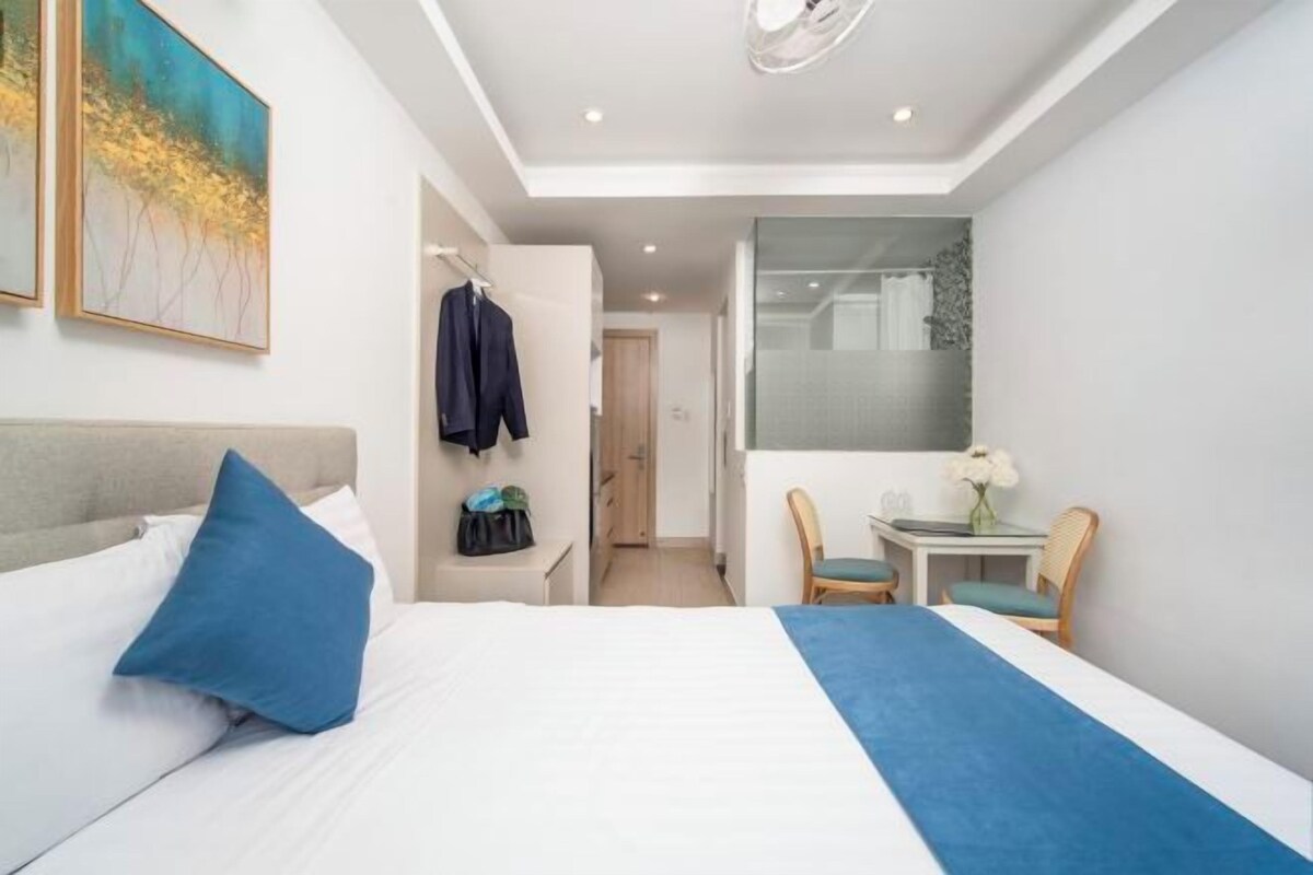 Quarto de hotel moderno com uma cama grande, uma escrivaninha e uma cadeira. Há um cabideiro com um casaco e uma bolsa, e um quadro na parede.