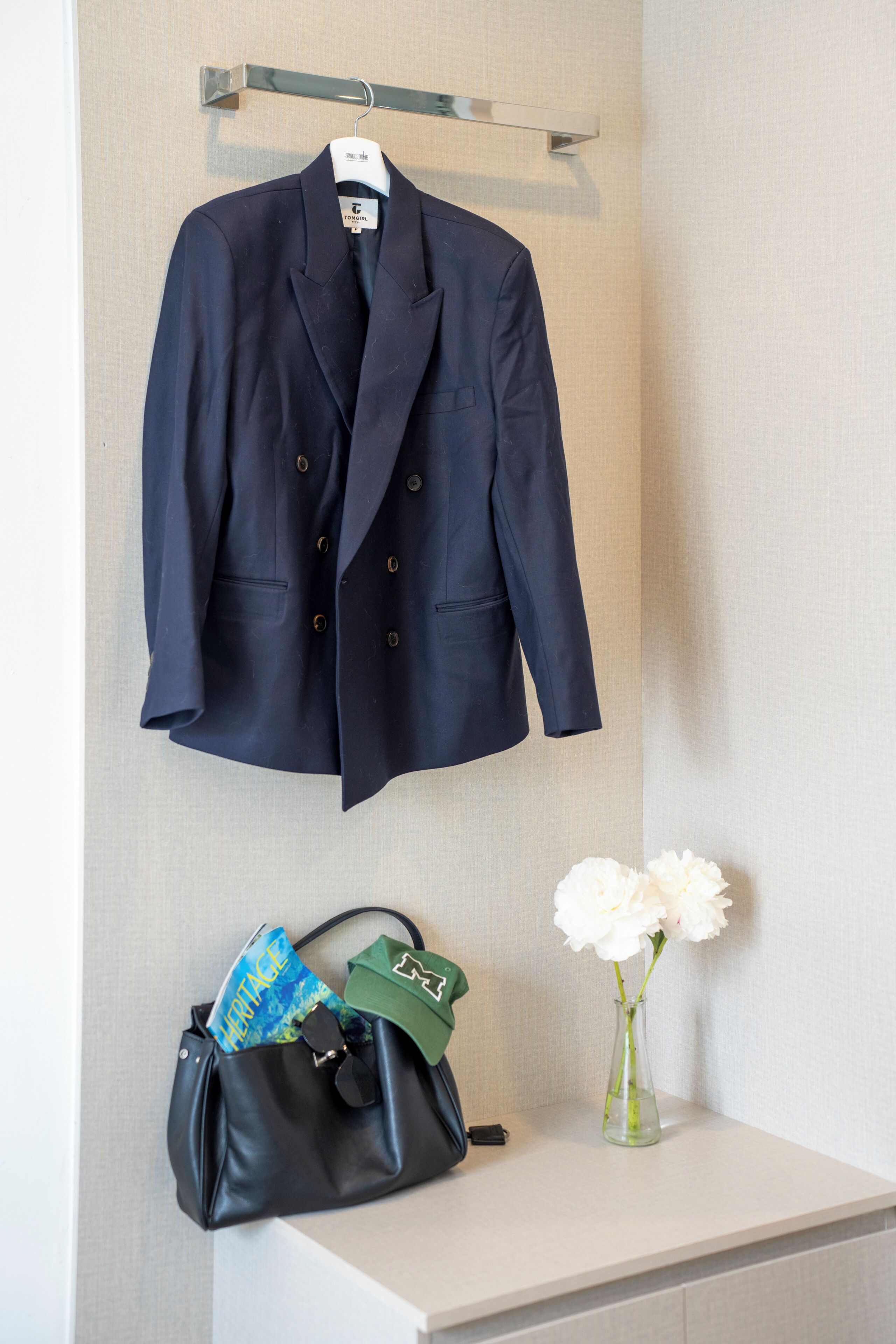 Un blazer blu scuro appeso a un attaccapanni a parete, una borsa in pelle nera con oggetti su un tavolo e un vaso con fiori bianchi.