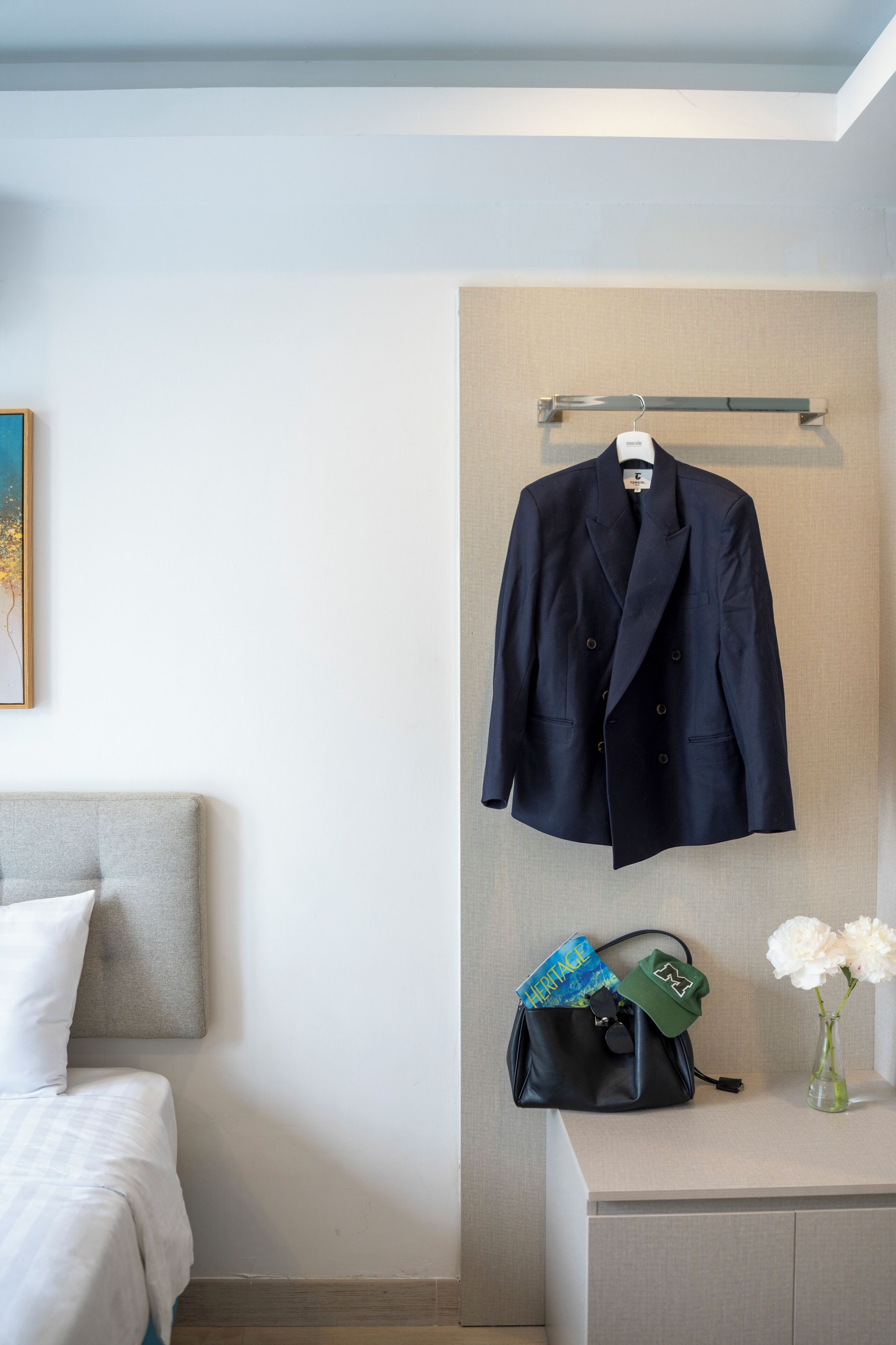 Habitación de hotel ordenada con una cama, una mesita de noche con un florero, un abrigo colgado y un bolso con una tapa verde.