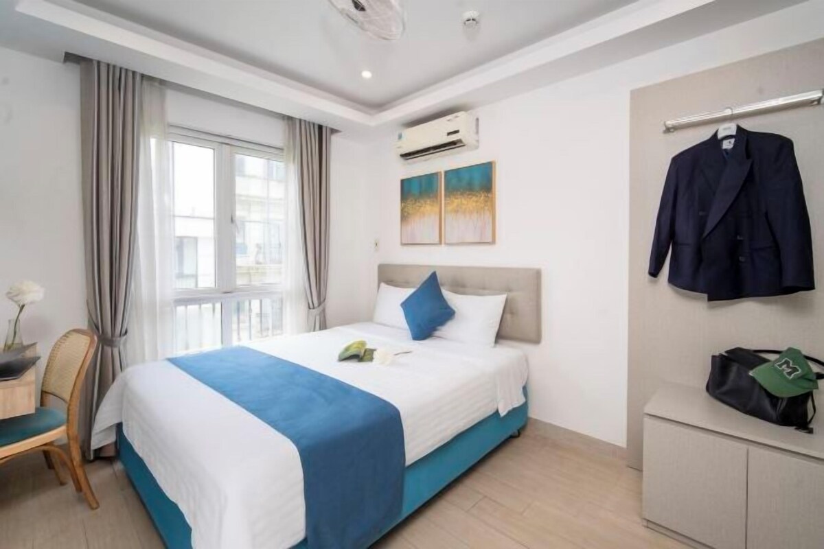 A hotel room with a bed, a desk, a chair, an air conditioning unit, and a coat rack with a jacket and a bag.