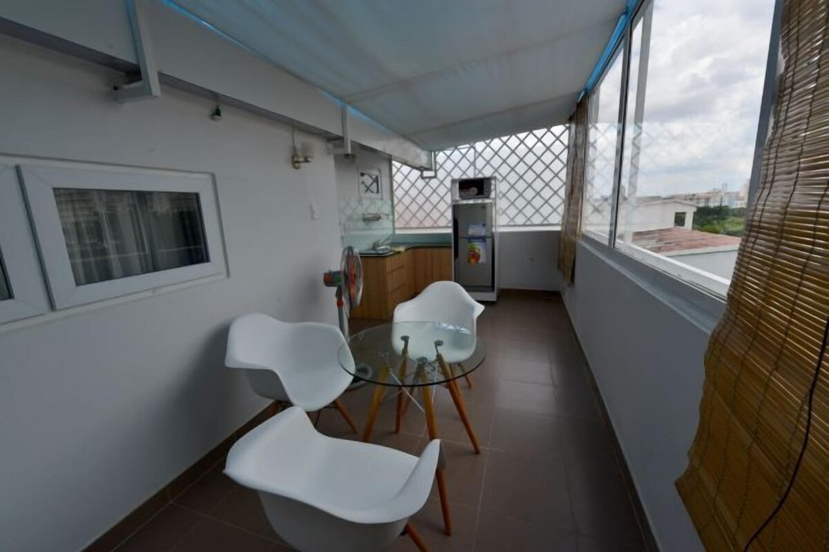 Uma varanda com mesa de vidro e cadeiras brancas, uma pequena cozinha e vista para a cidade.