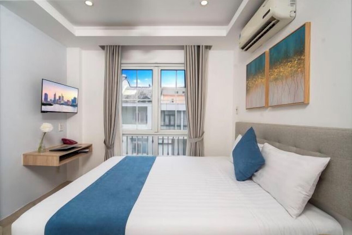 A hotel room with a bed, a mounted TV, a window with curtains, and artwork on the wall.