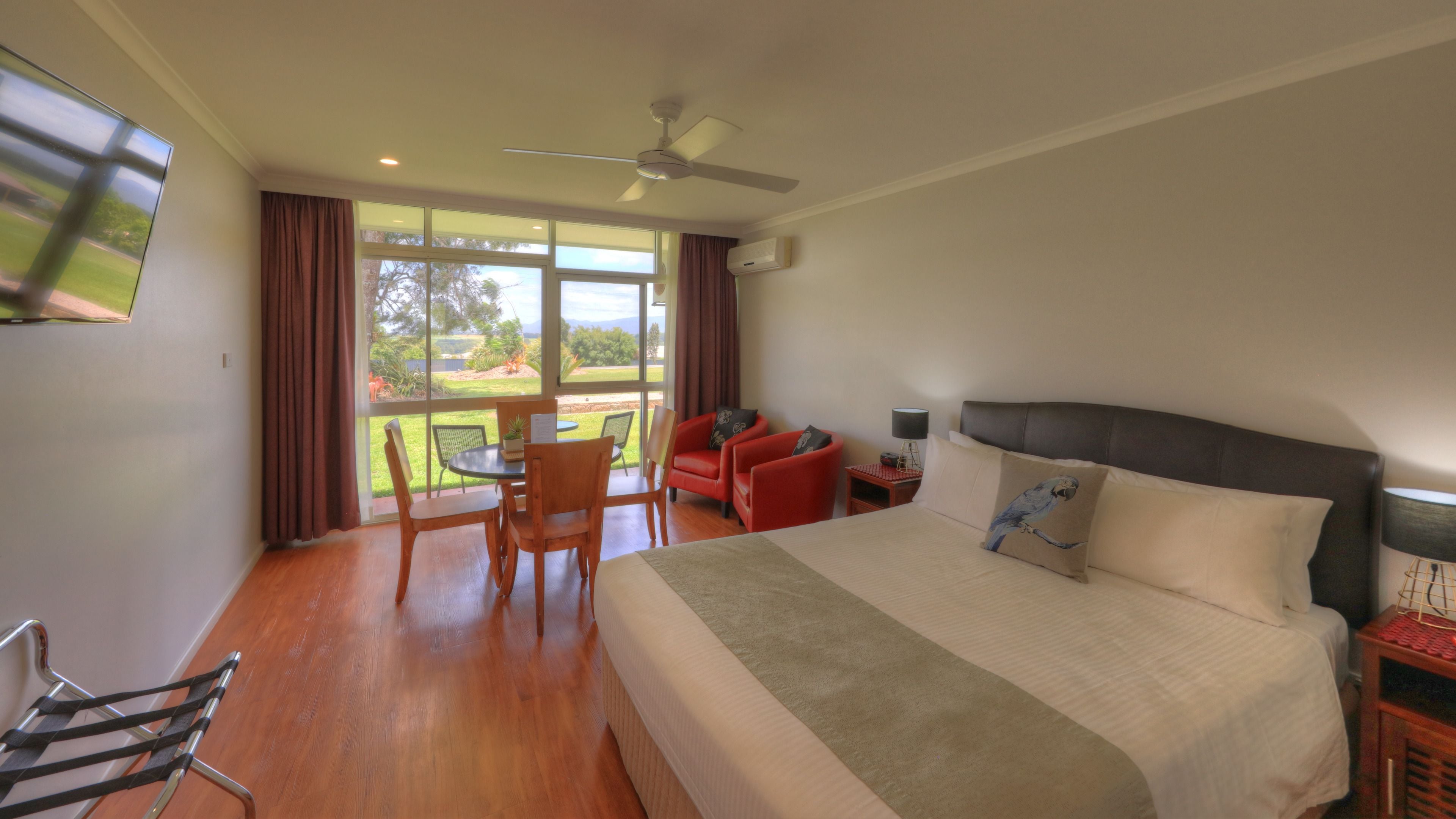 A hotel room with a large bed, a dining table with chairs, a red armchair, a ceiling fan, and a view of the outdoors.