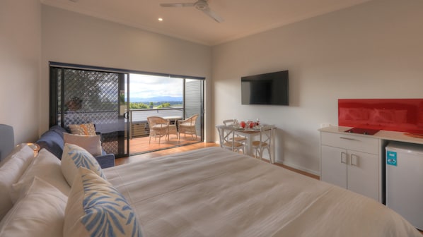 A bedroom with a large bed, a TV mounted on the wall, a small table with chairs, and a balcony with wicker furniture. - Atherton Motel (Atherton)