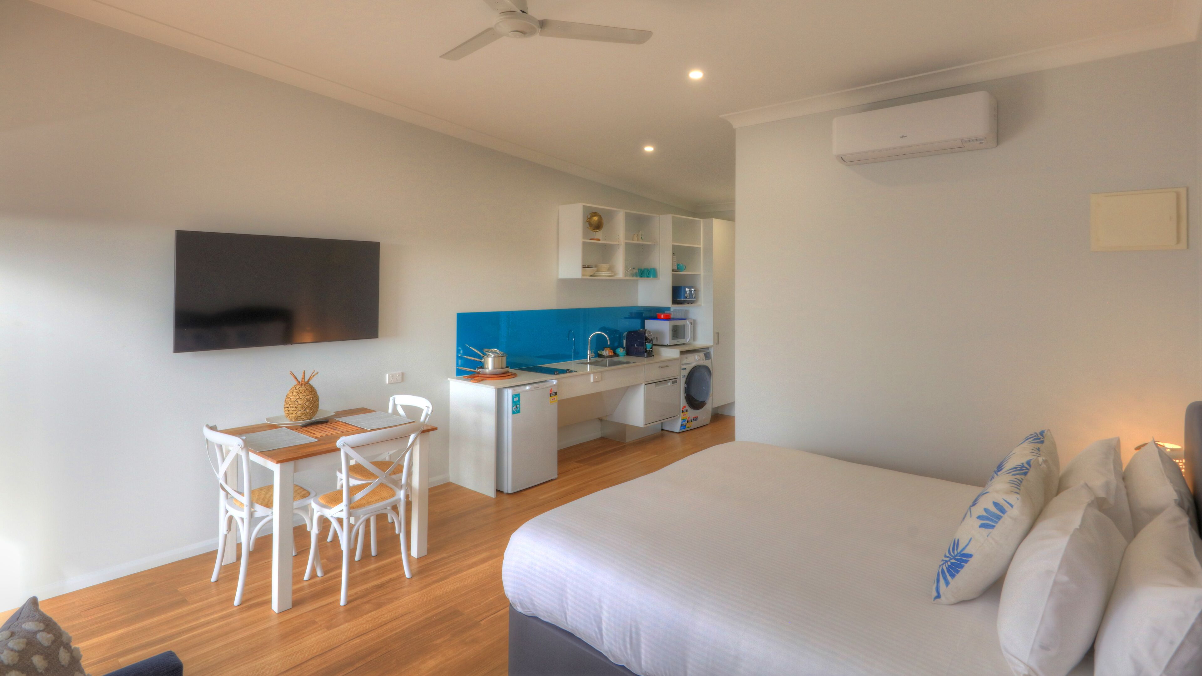 A compact hotel room with a bed, a desk with chairs, a kitchenette, and a TV mounted on the wall.