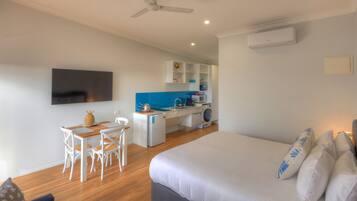 A compact hotel room with a bed, a desk with chairs, a kitchenette, and a TV mounted on the wall.