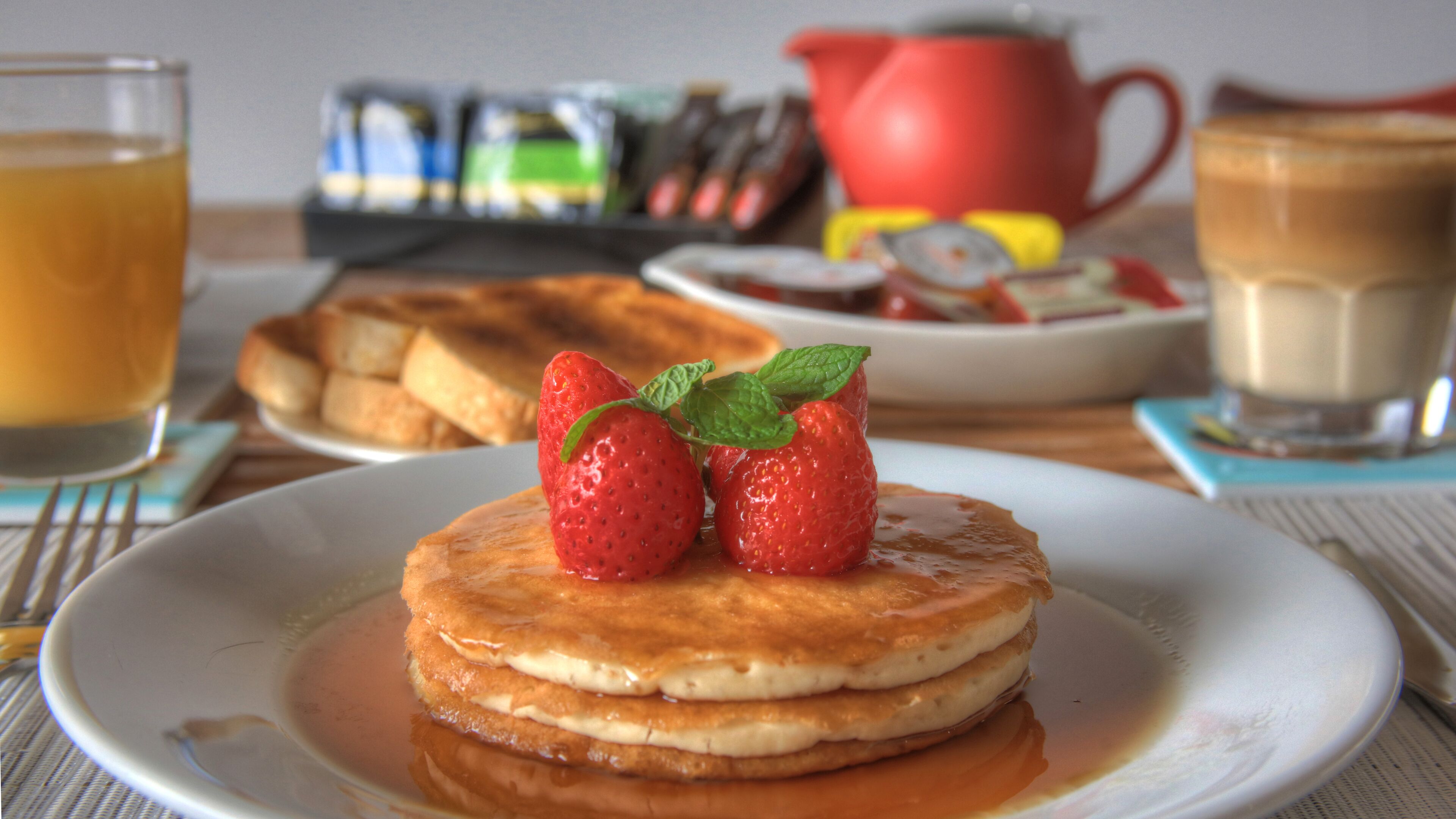 A breakfast plate with pancakes topped with strawberries and syrup, a glass of orange juice, and a red teapot in the background.