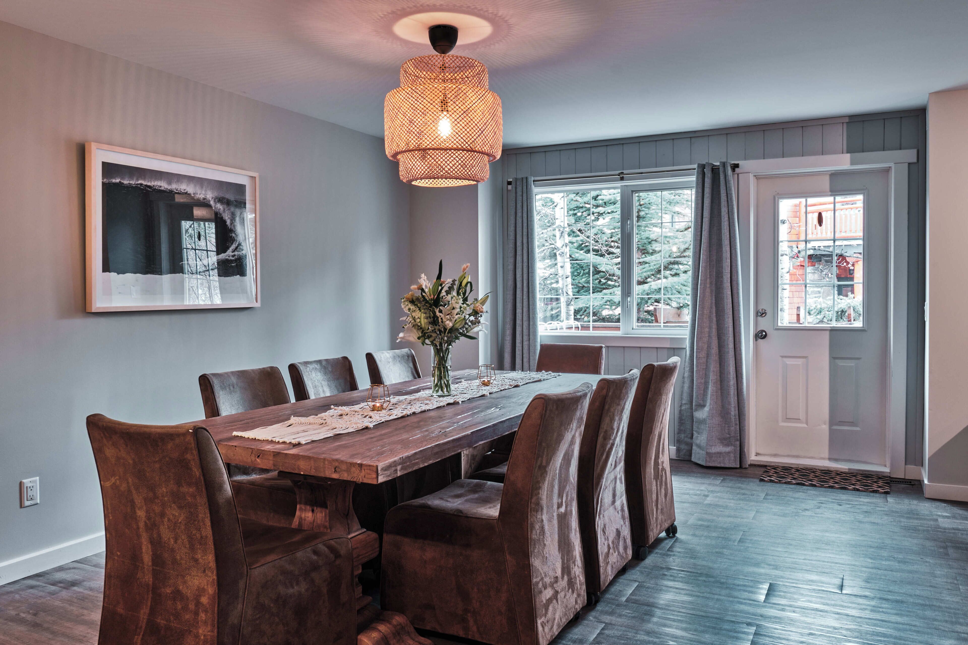 A dining room with a wooden table, leather chairs, a hanging light fixture, a framed picture on the wall, and a door with a view of trees.