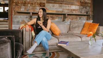 Una persona sentada en un sofá con una bebida, en una habitación con paredes de madera y una mesa de centro con un libro y flores.