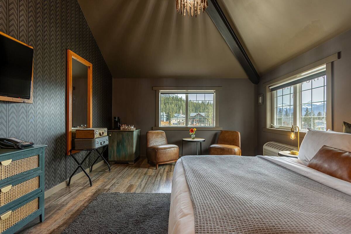 A cozy bedroom with a large bed, a desk with a telephone, a chair, a small table with a vase, and a window with a view of trees and mountains.