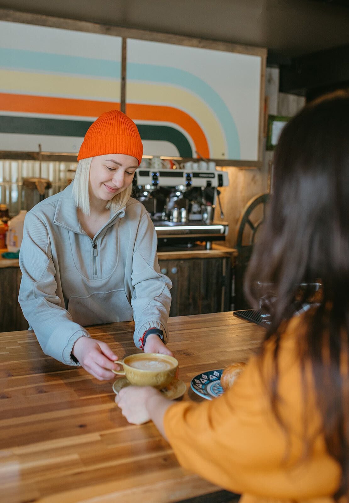A person serving coffee to another person at a wooden table in a cafe with a colorful wall in the background.
