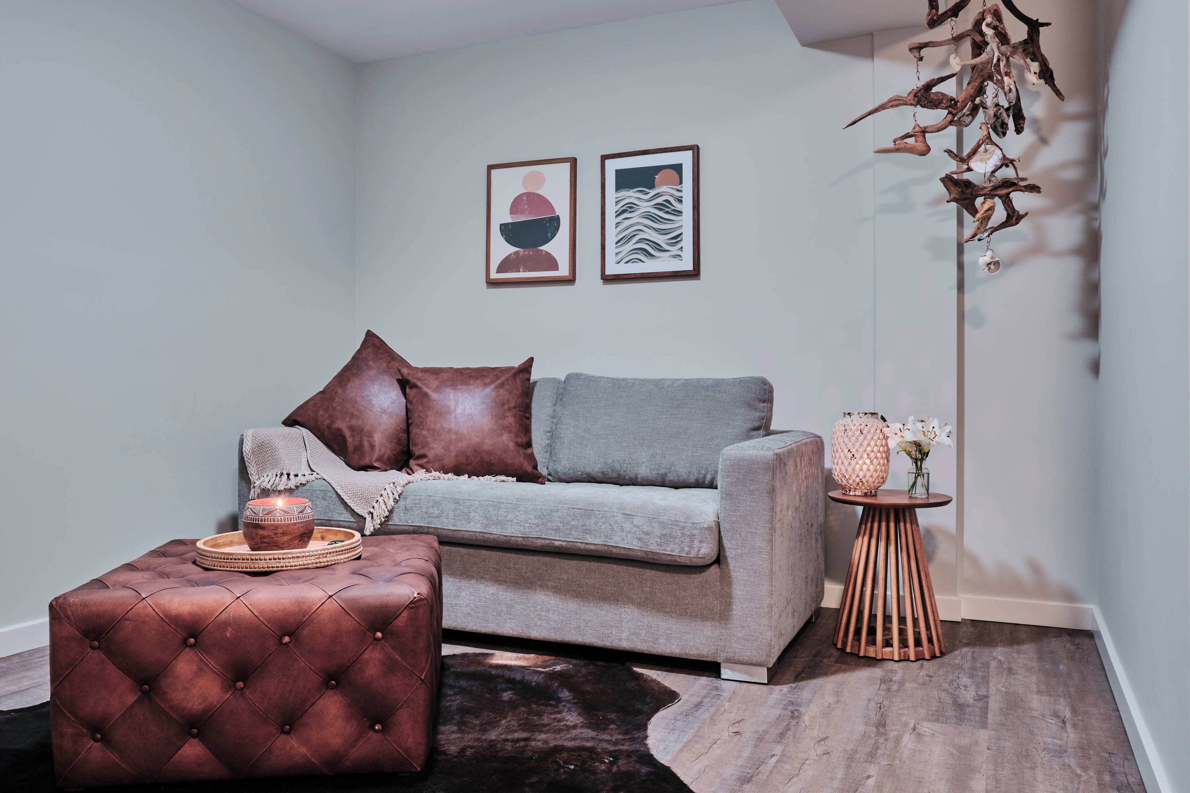 Una sala de estar moderna con un sofá gris, cojines marrón y gris, un otoman marrón y una pequeña mesita de madera con un jarrón y flores.