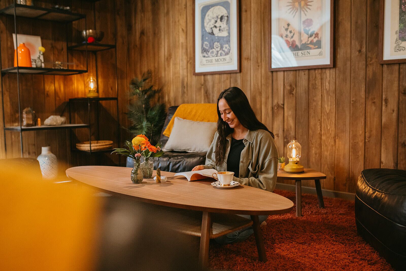 Una habitación acogedora con paredes de madera, una mesa redonda de madera con un libro, una taza y jarrones de flores. Hay carteles enmarcados en la pared y una pequeña mesa con una lámpara.