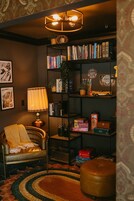 Quarto aconchegante com poltrona de couro, estante cheia de livros e jogos de tabuleiro, abajur e tapete estampado.