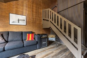 Una sala de estar moderna con un sofá gris, un cojín a rayas rojo y amarillo, un amplificador Marshall y una maleta antigua.