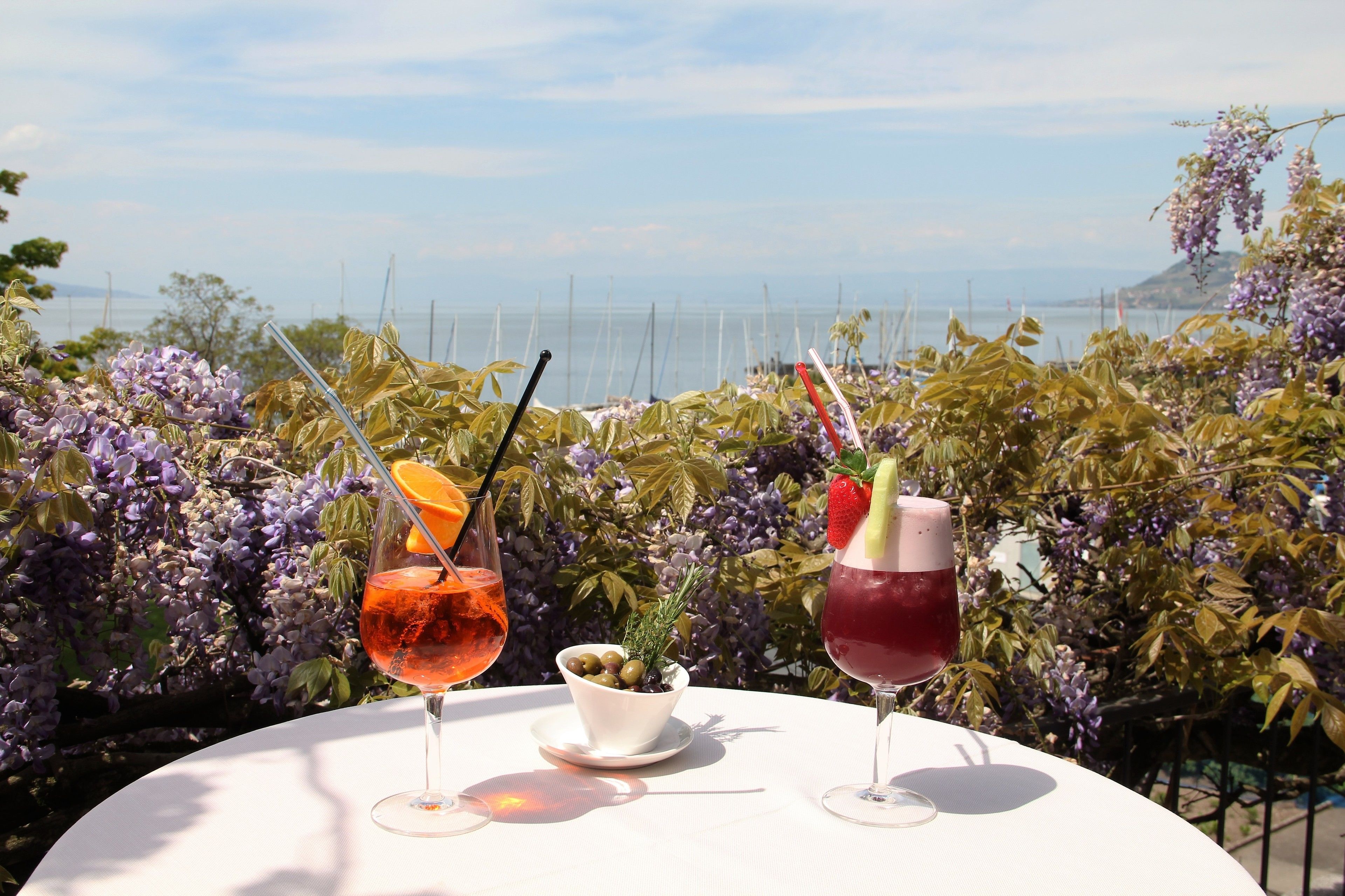 Una mesa con dos bebidas y un cuenco pequeño de aceitunas, frente a un fondo de flores moradas y vistas al mar.