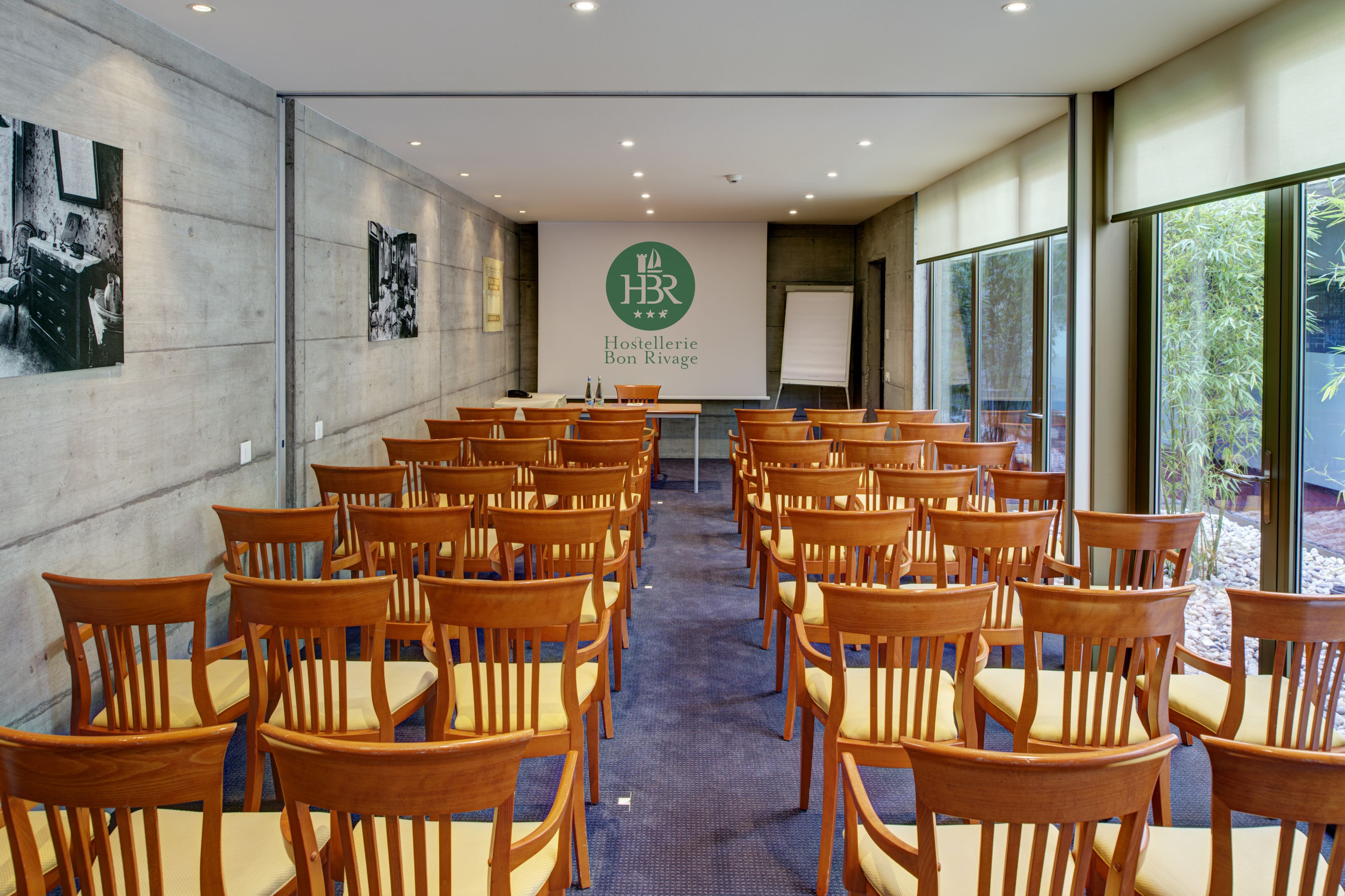 Una sala de conferencias con filas de sillas de madera y una pantalla grande que muestra el logotipo de Hostellerie Bon Rivage.