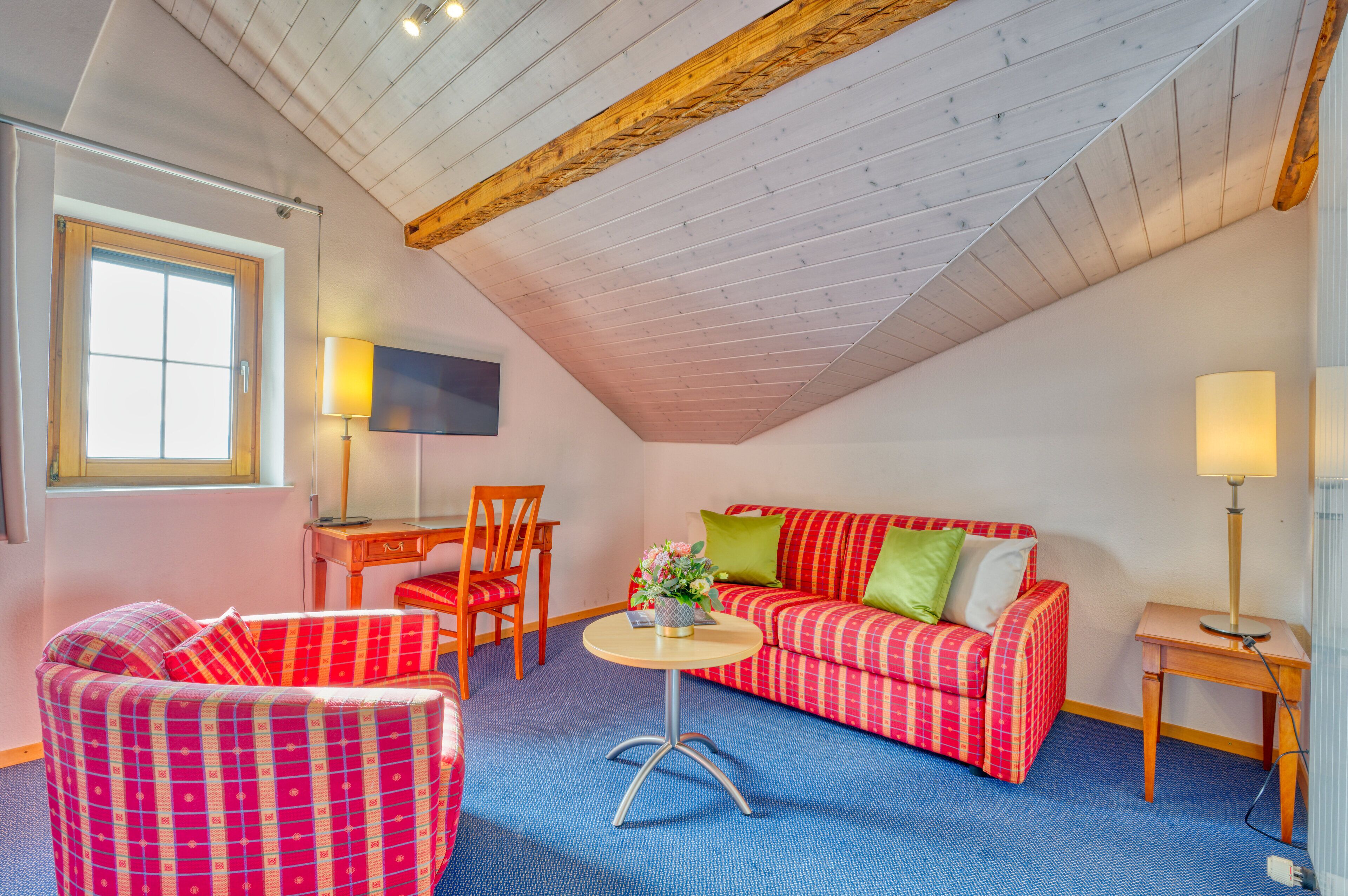 A cozy room with a sloped ceiling, a red and white checkered sofa, a matching armchair, a small round table with a lamp, and a TV on the wall.