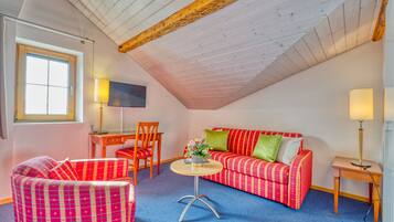 A cozy room with a sloped ceiling, a red and white checkered sofa, a matching armchair, a small round table with a lamp, and a TV on the wall.
