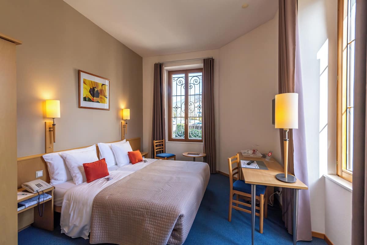Ein Hotelzimmer mit einem großen Bett, einem Schreibtisch, einem Stuhl, einem kleinen Tisch, einer Lampe, einem Telefon und einem Fenster mit Vorhängen.